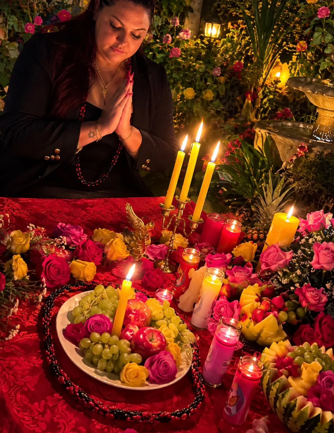 Alice em oração diante de velas amarelas, rosas, frutas e um colar de contas vermelhas e pretas, em jardim iluminado
