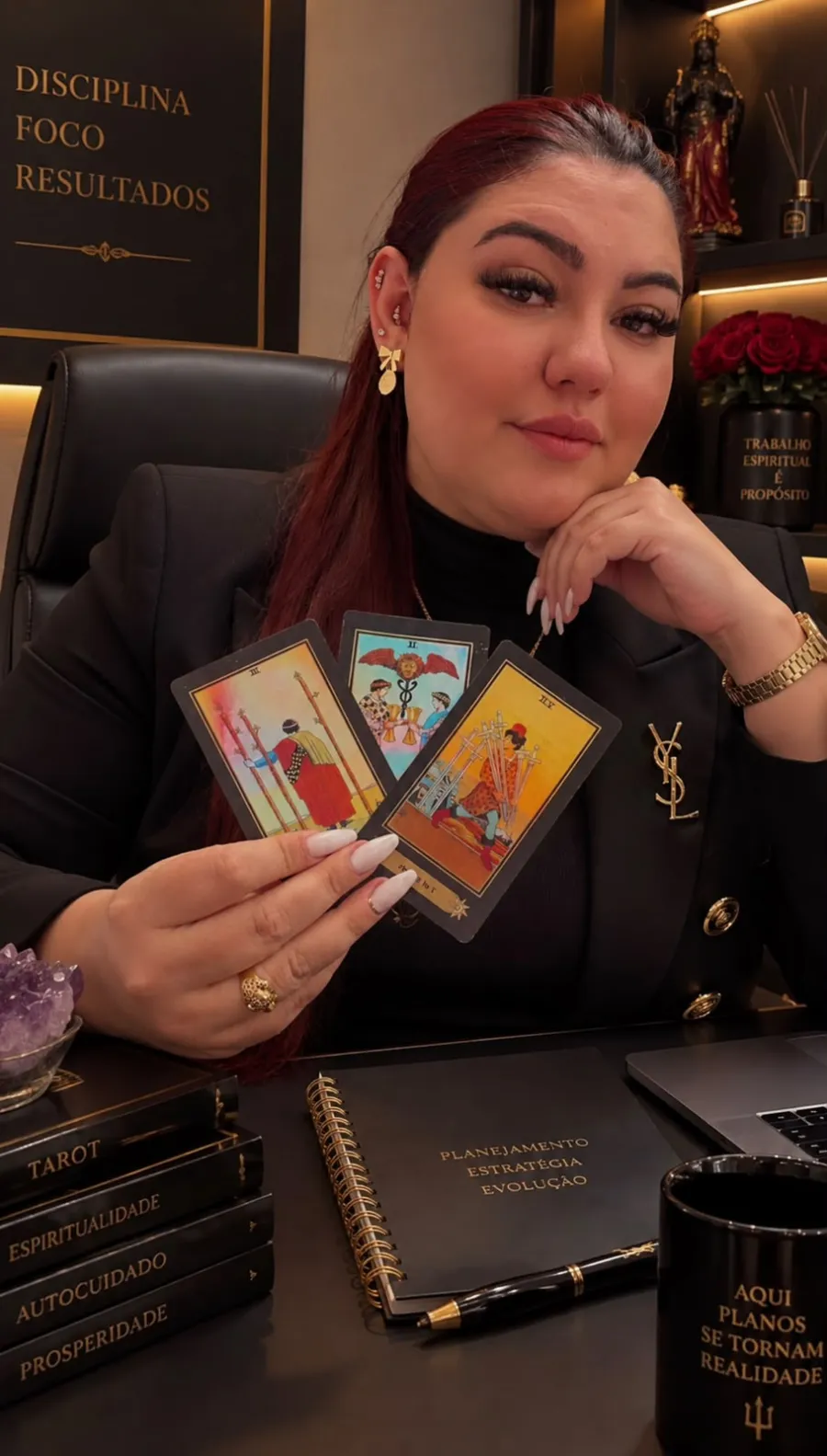 Alice segurando três cartas de tarô em seu consultório, com livros e uma ametista sobre a mesa